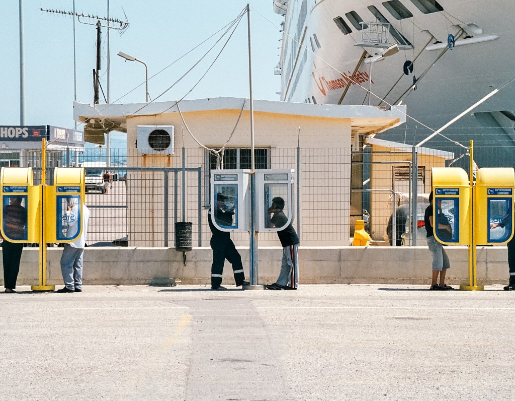People using yellow public telephones outside a small beige building, a ship behind.