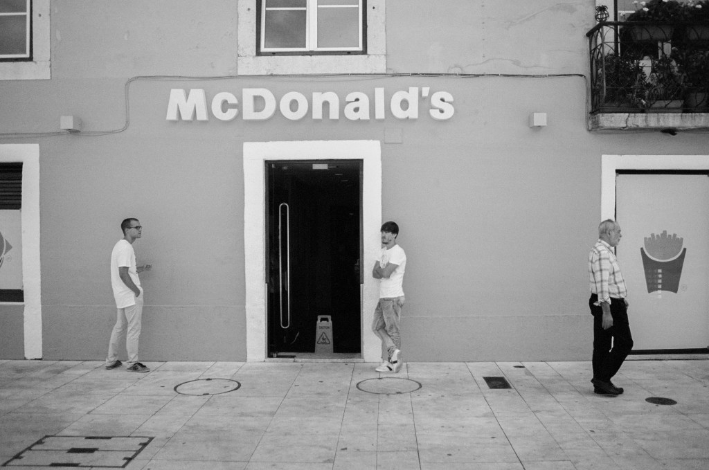 Black and white. Three men standing apart outside a McDonald's doorway.
