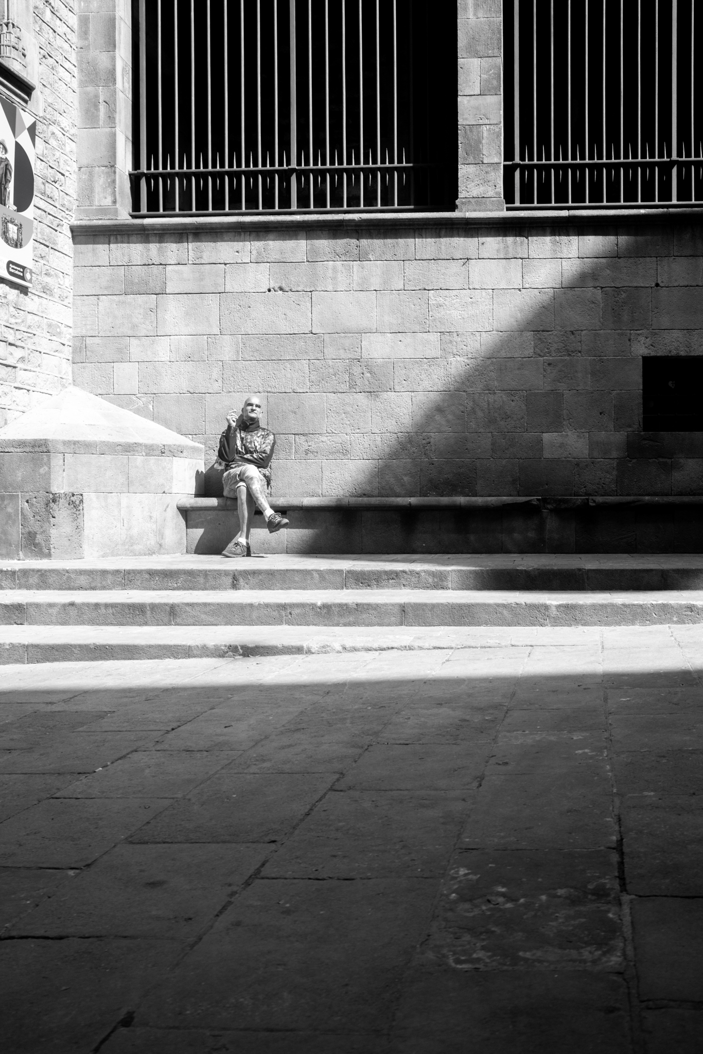 Black and white. A person sitting on stone steps in a patch of sunlight below barred windows.