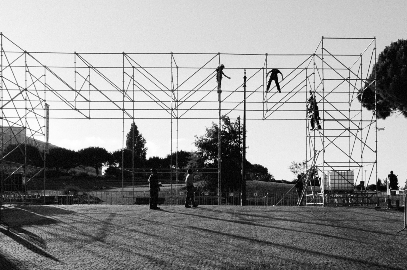 Black and white. Workers climbing a scaffold frame while others stand below.