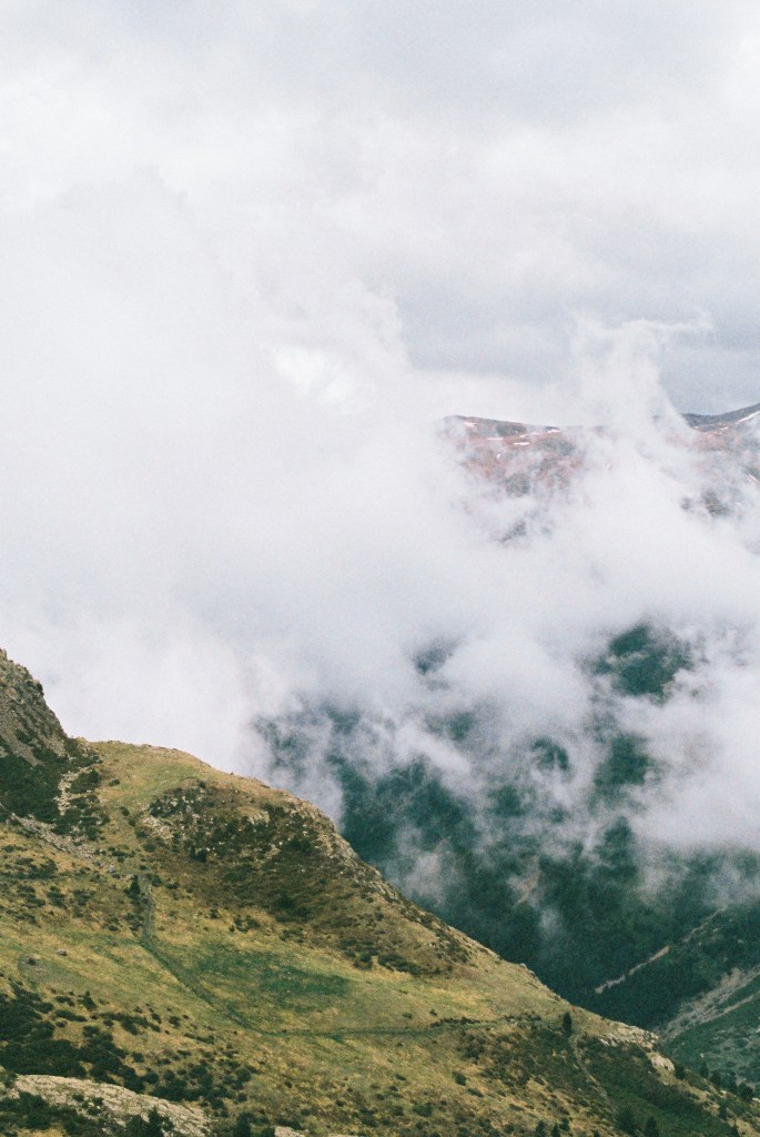 Cloud rising out of a valley between a grassy slope and a distant ridge.