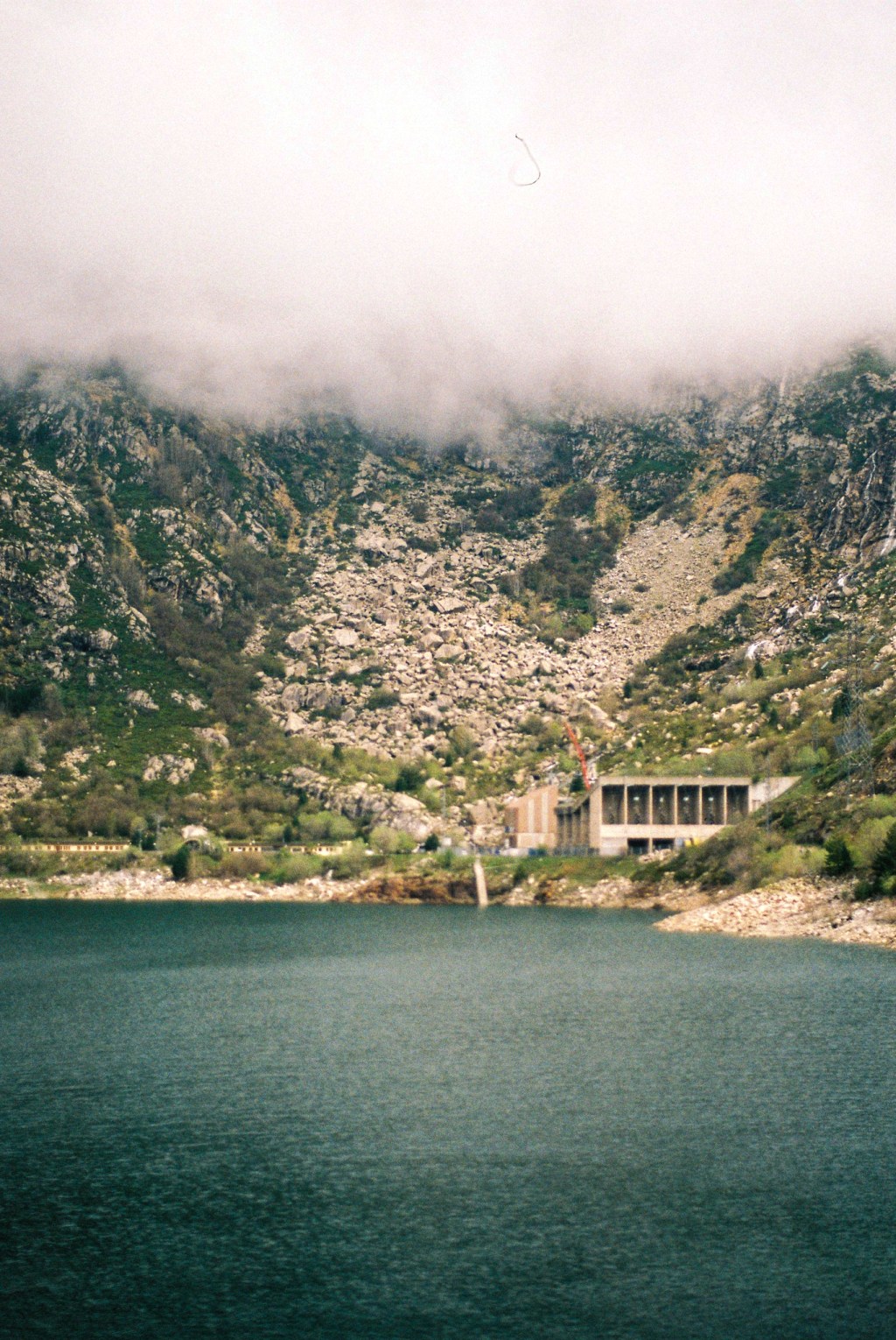 A dam and a green reservoir beneath a scree slope, the mountain above lost in cloud.