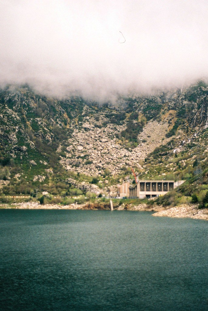 A dam and a green reservoir beneath a scree slope, the mountain above lost in cloud.