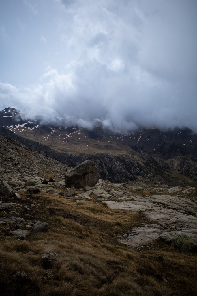 Low cloud covering a mountain ridge above a boulder-strewn slope of brown grass.